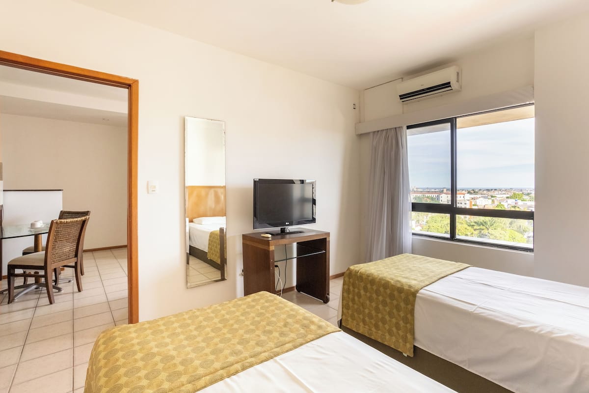 A hotel room with two beds, a desk with a chair, a television, and a large window with a city view.