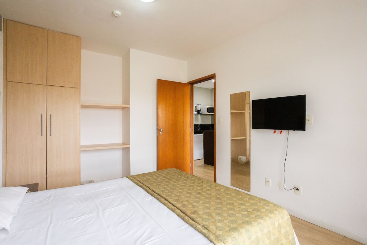 A bedroom with a bed, wooden wardrobe, a television mounted on the wall, and a kitchen area visible through an open door.