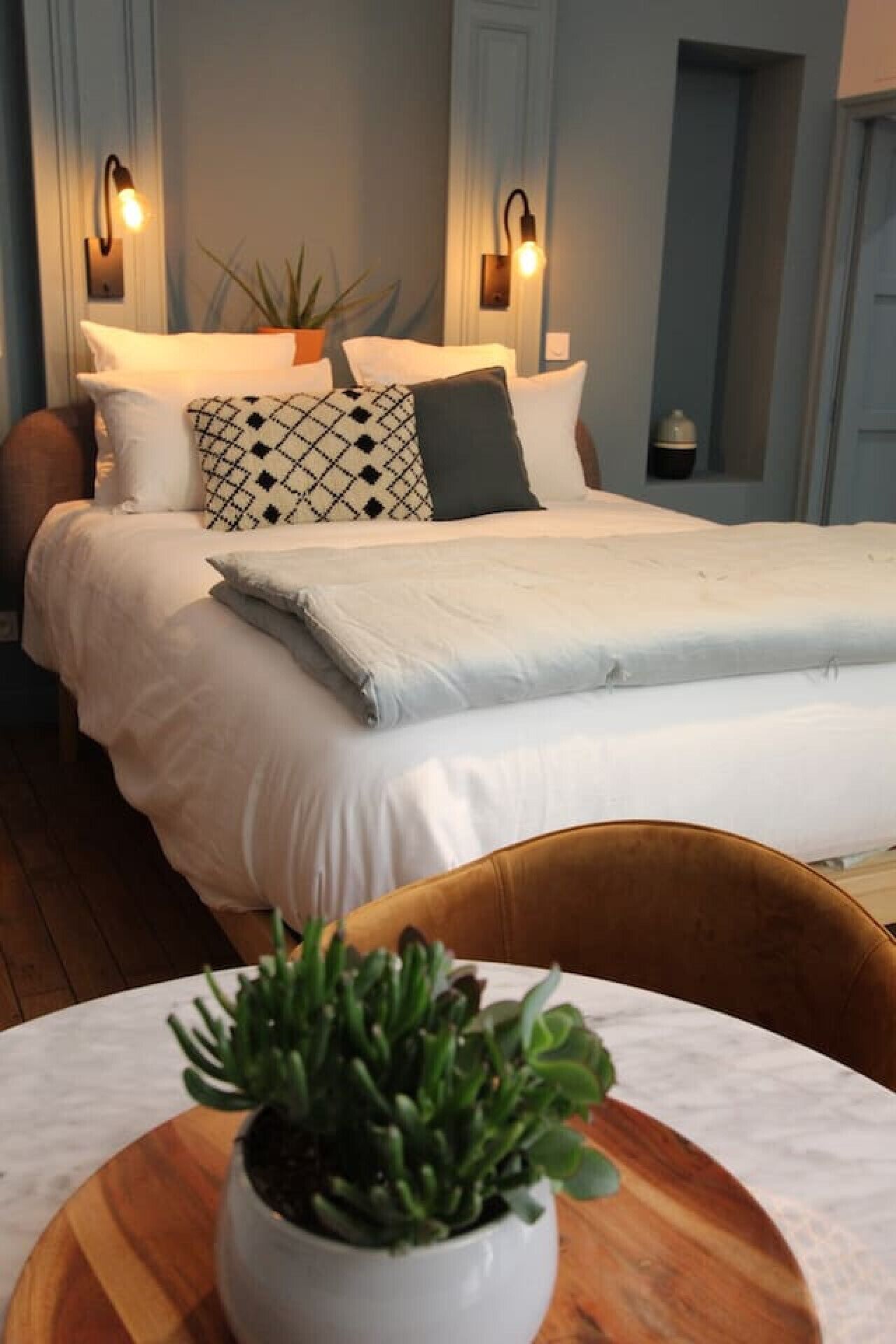 A bedroom with a bed, two bedside lamps, a potted plant, and a wooden chair with a table.
