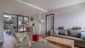 Una sala de estar moderna con sofá, reposapiés y mecedora. Se ve un comedor con mesa y sillas a través de una puerta de vidrio.