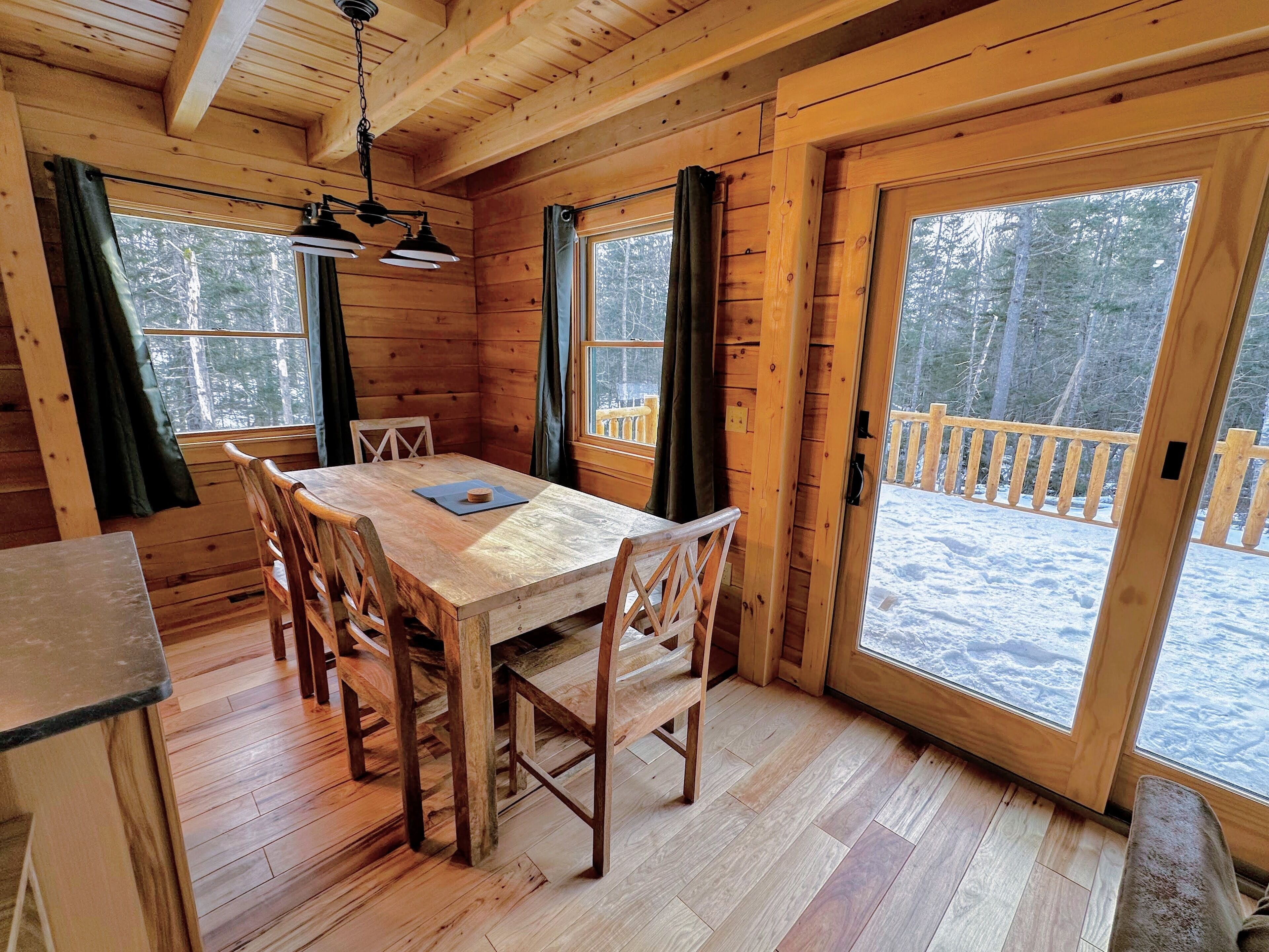 L’intérieur d’un chalet en bois, avec une table à manger, des chaises et une fenêtre donnant sur une terrasse enneigée.