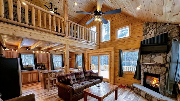 Uma cabana espaçosa com interior em madeira, lareira de pedra, área de refeições e uma sala de estar aconchegante com sofá e mesa de centro.