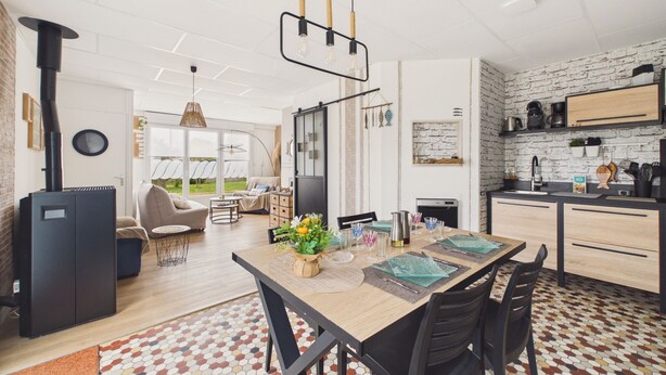 A modern kitchen with a brick wall backsplash, a dining area with a wooden table, and a visible outdoor view through large windows. - Flat for 4 with large garden (Préfailles)