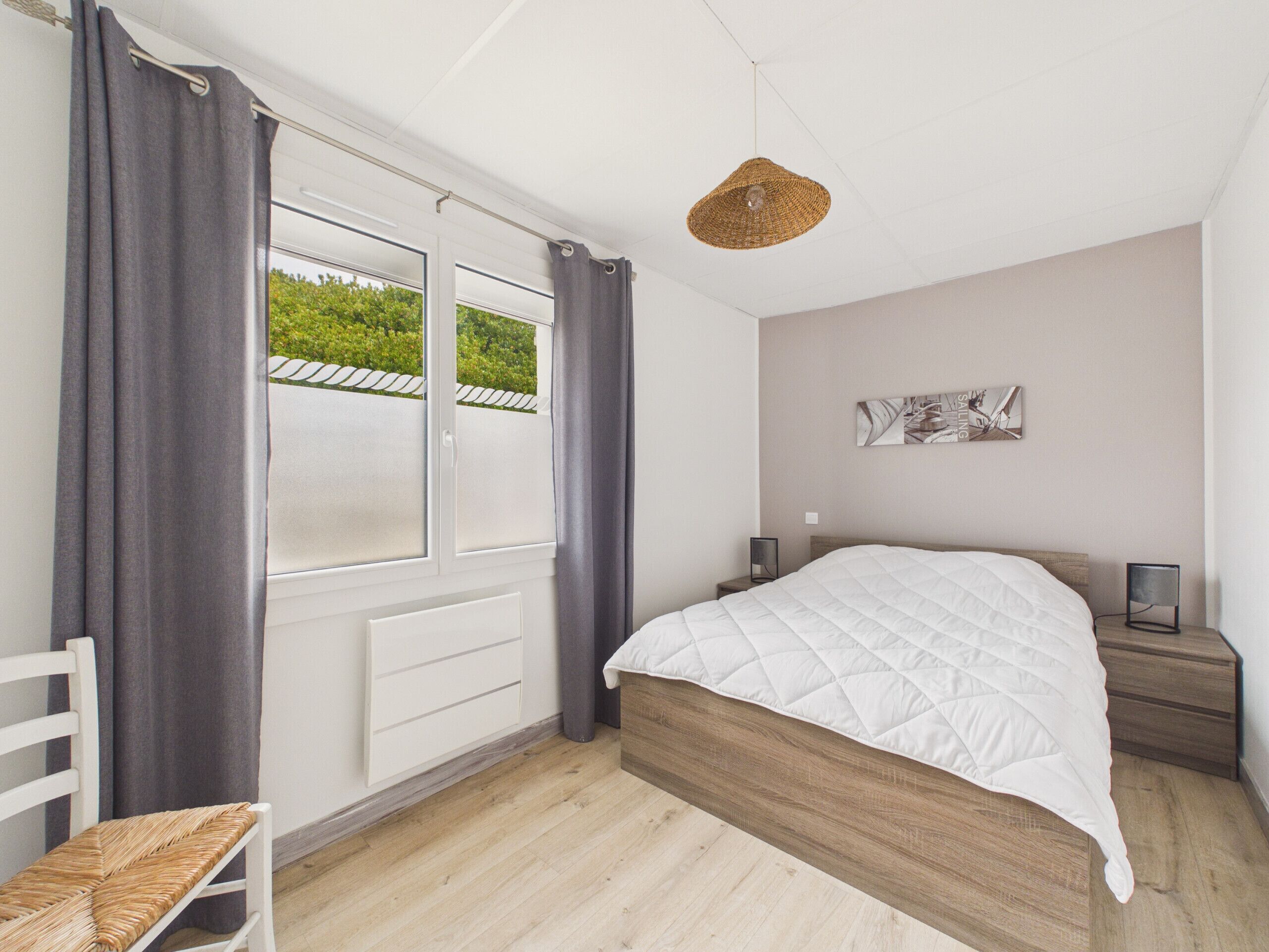 Un dormitorio con cama, mesitas de noche, una silla, una ventana con cortinas, una lámpara colgante y una obra de arte en la pared.