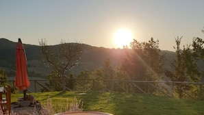 Mão segurando uma caneca com vista para um campo gramado e um pôr do sol ao fundo.