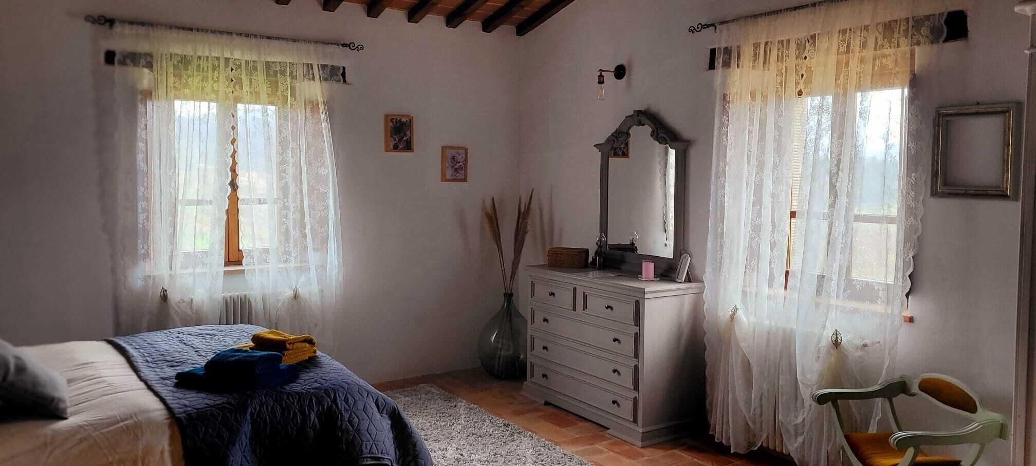 A bedroom with a bed, dresser, and two windows with sheer curtains.