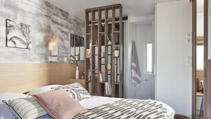 A modern bedroom with a large bed, a wooden partition, a wall-mounted mirror, and a framed picture. - Camping Pradelongue (Moustajon)