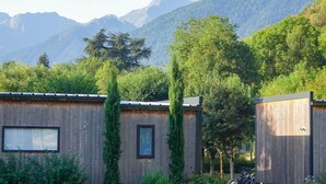 A rustic cabin with a wooden exterior, a window, and a door. It is surrounded by lush greenery and mountains in the background. - Camping Pradelongue (Moustajon)