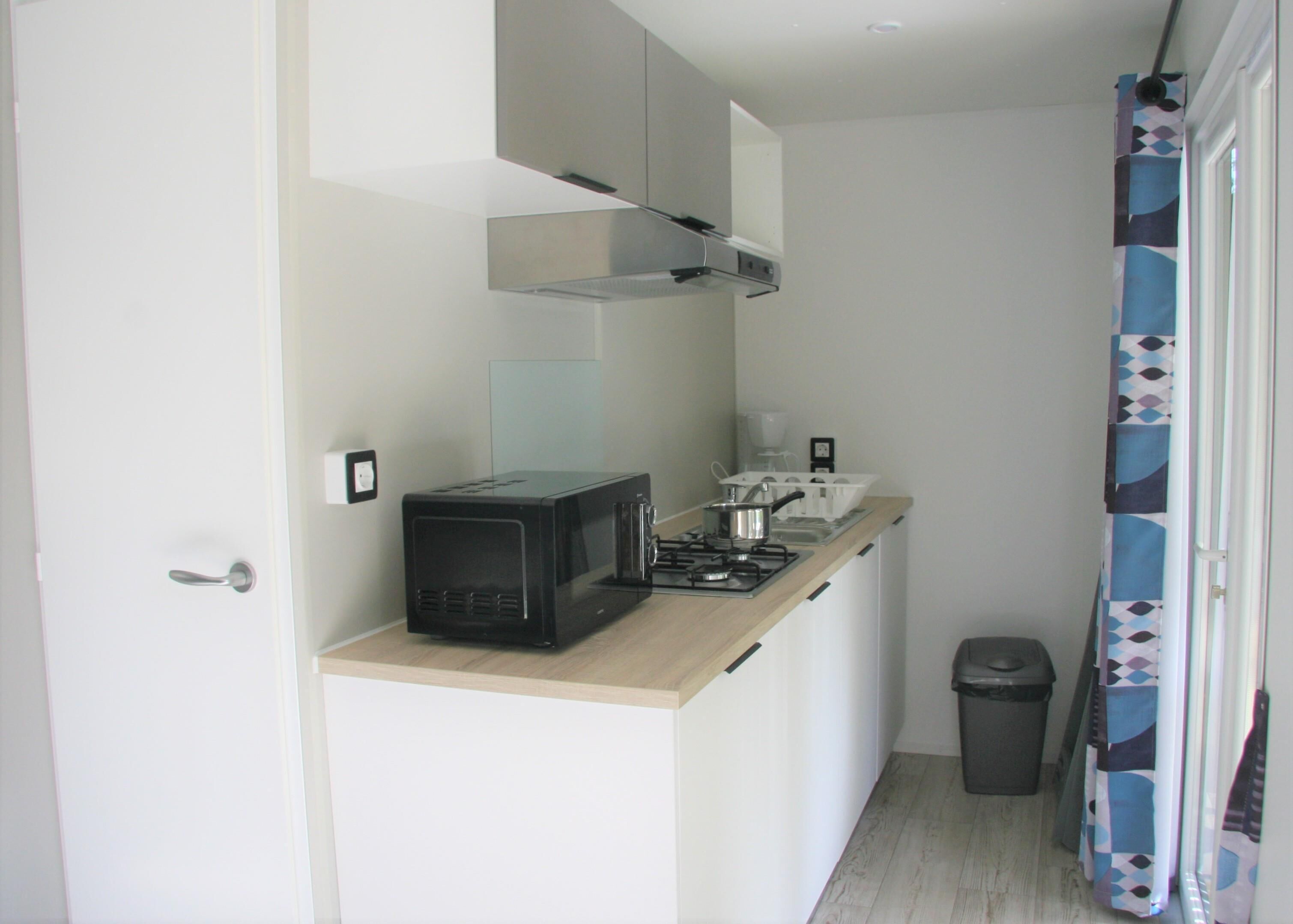 Cocina compacta con microondas, estufa y lavamanos. Incluye un cubo de basura y una ventana con cortina.