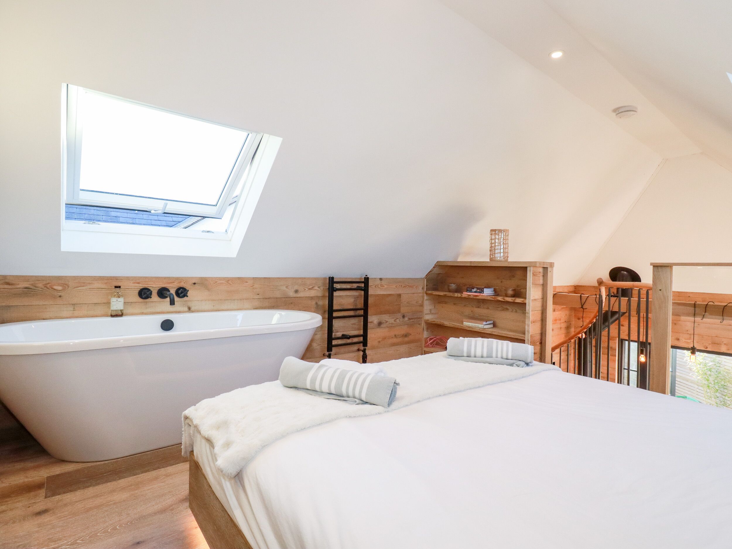 A modern bedroom with a skylight, a large bathtub, wooden flooring, and a staircase.