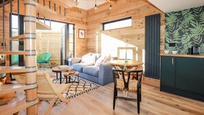 A spacious living area with a wooden staircase, a blue sofa, a dining table, and a kitchen area with a green leaf-patterned backsplash. - Little Beach Cabin (Newquay)