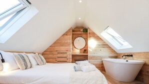 A bedroom with a large bed, a wooden headboard, a skylight, a round mirror, and a freestanding bathtub. - Little Beach Cabin (Newquay)
