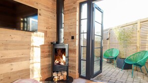 A cozy room with a wood-burning stove, a large sliding glass door leading to an outdoor patio with chairs and a bamboo plant. - Little Beach Cabin (Newquay)