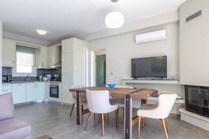 Una cocina moderna con armarios blancos, una mesa de comedor con sillas, un televisor montado sobre la chimenea y una pequeña isla de cocina con un hervidor.