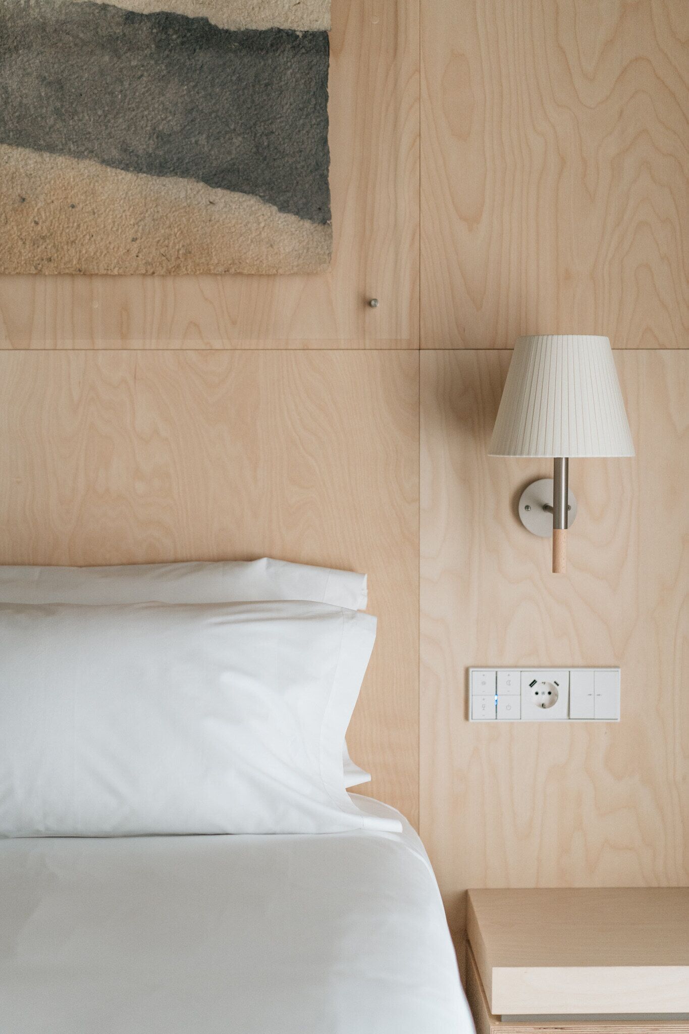Een net opgemaakt bed met witte lakens, een wandlamp en een stopcontact op een houten wand.