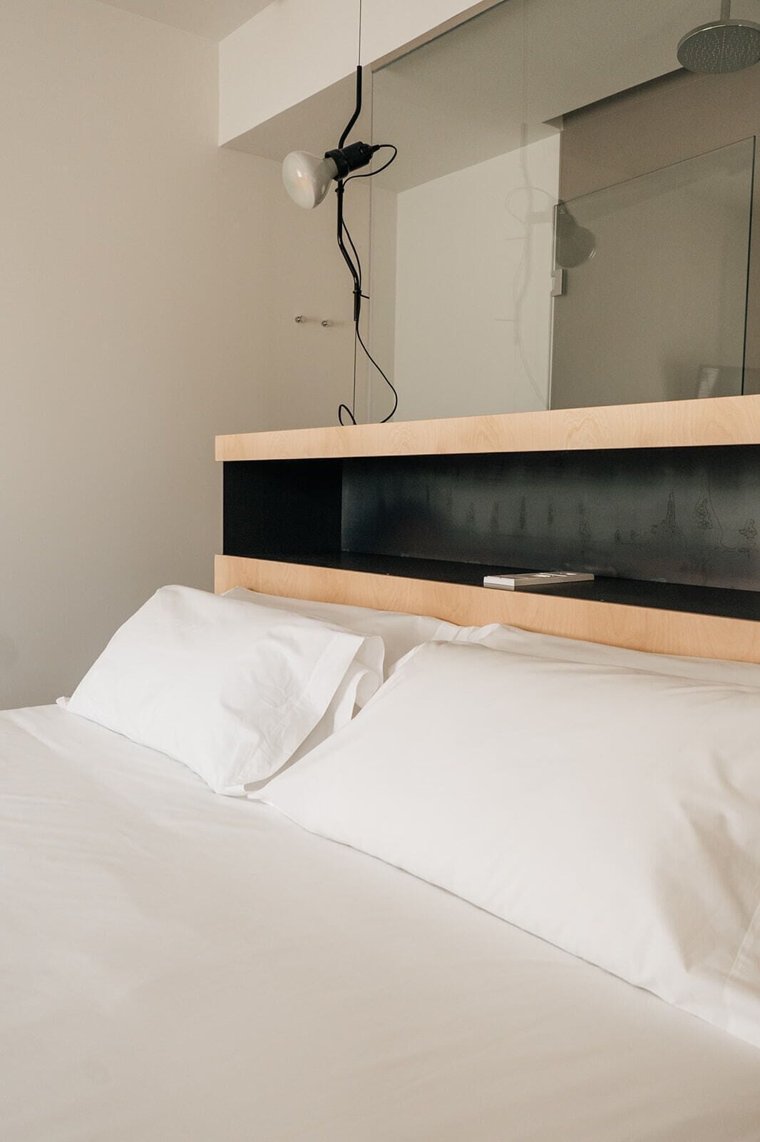 A modern bedroom with a bed, white linens, a wooden headboard, a mirror, and a wall-mounted light fixture.