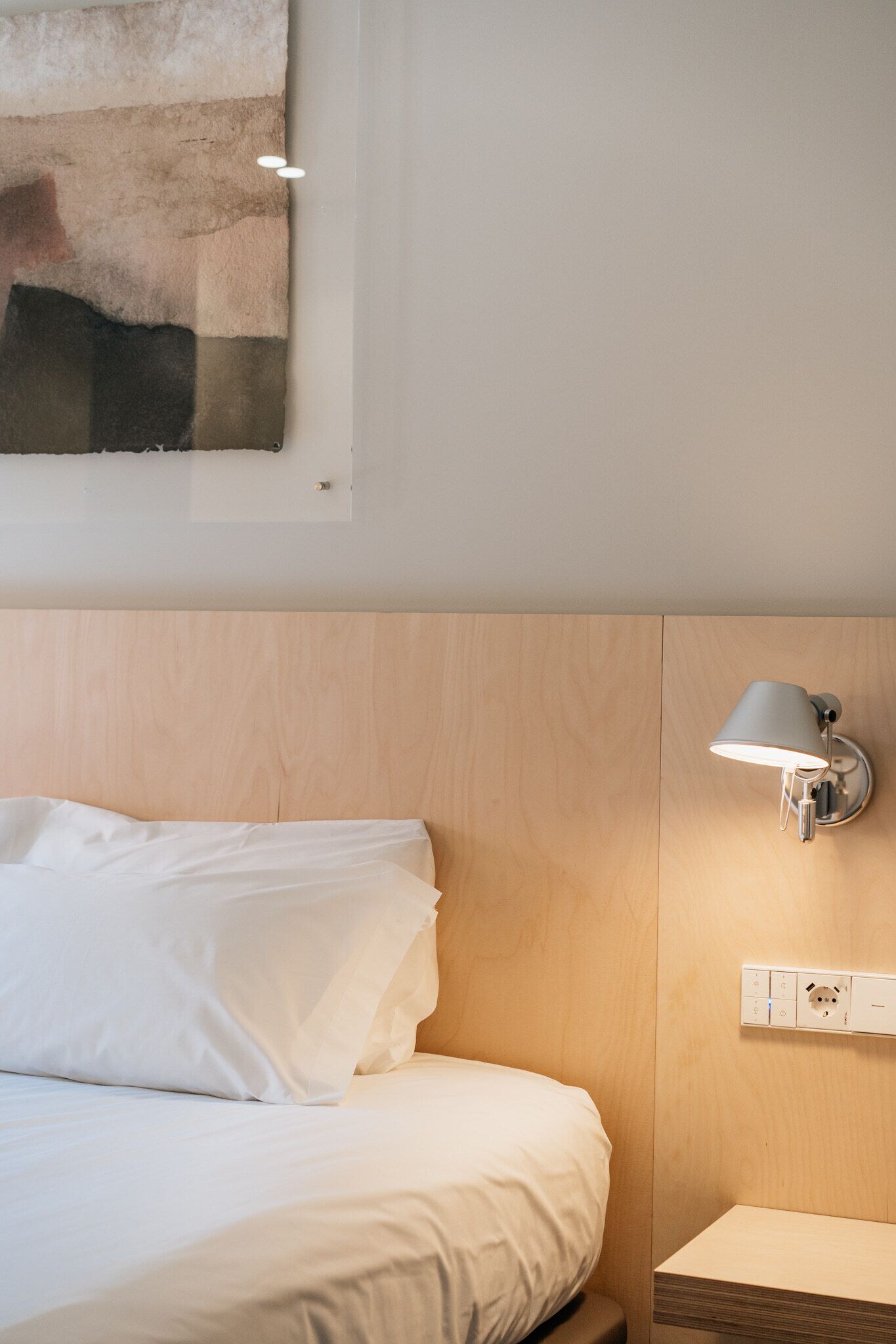 A hotel room with a wooden headboard, a white bed, a wall-mounted lamp, and a power outlet.