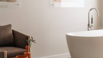 A modern bathroom with a freestanding bathtub, a wooden table with a book and a vase, and a brown sofa.
