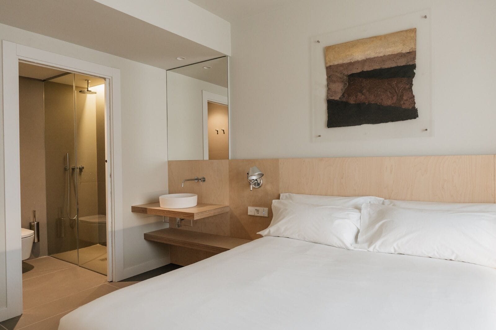 Ein modernes Hotelzimmer mit einem großen Bett, einem Nachttisch mit Waschbecken, einer Duschkabine und einem an der Wand hängenden Kunstwerk.