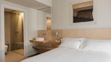 Ein modernes Hotelzimmer mit einem großen Bett, einem Nachttisch mit Waschbecken, einer Duschkabine und einem an der Wand hängenden Kunstwerk.