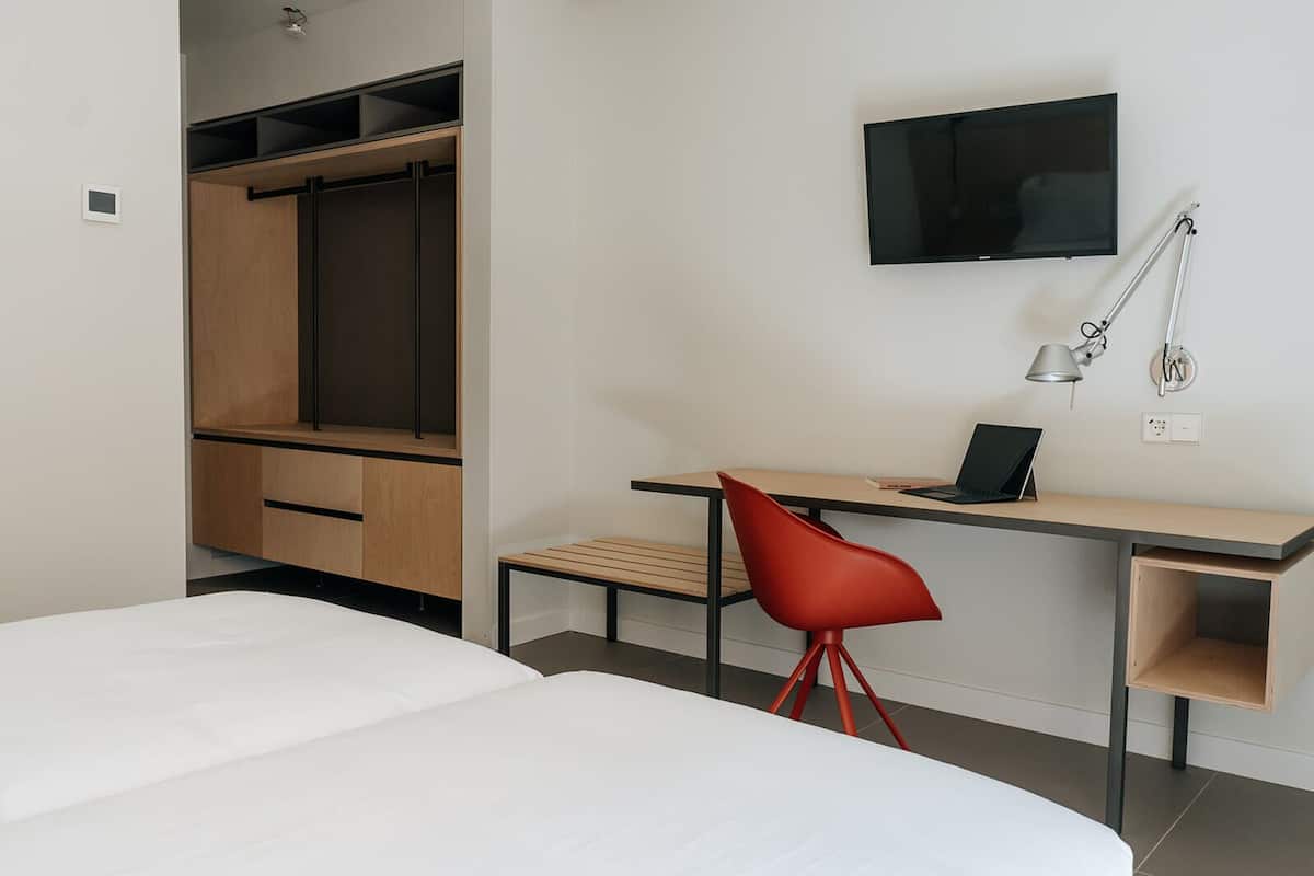 A modern hotel room with a bed, a desk with a laptop, a red chair, a TV, and a wardrobe.