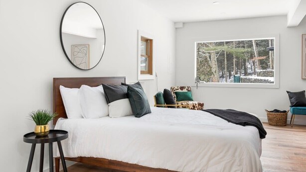 Une chambre moderne avec un grand lit, un miroir rond, une fenêtre donnant sur la verdure et une petite table d’appoint agrémentée d’une plante.