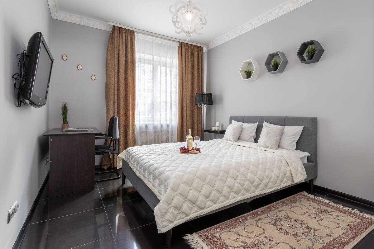 A modern bedroom with a bed, a desk, a chair, a TV, and decorative plants.
