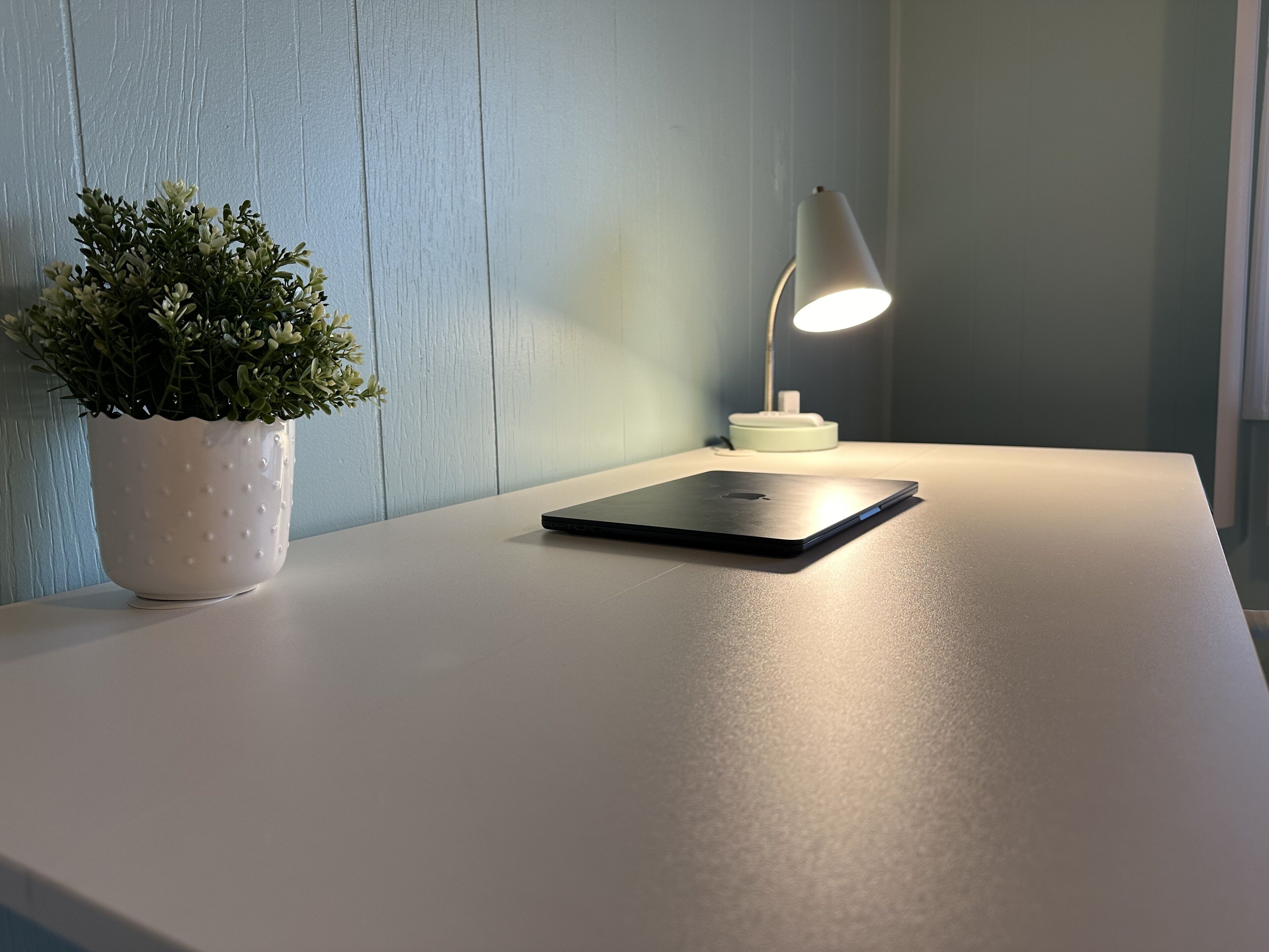 En bärbar dator står på ett vitt bord tillsammans med en krukväxt och en bordslampa.
