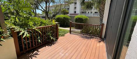 A wooden deck with a railing, surrounded by greenery and a building in the background.