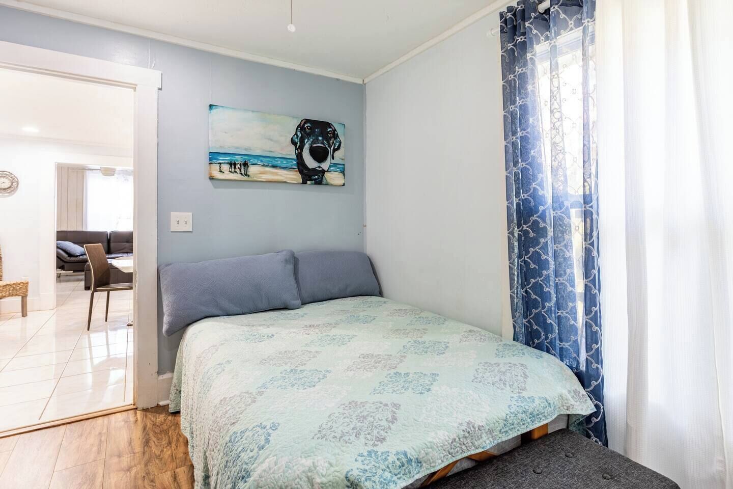 A bedroom with a bed, a painting of a dog, and a window with curtains.