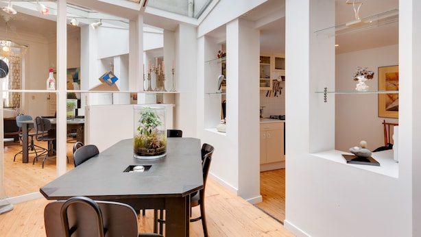 Un intérieur moderne avec un coin repas comprenant une table à plateau en verre, un arrangement floral dans un pot et une vue sur la cuisine et l’espace de vie.
