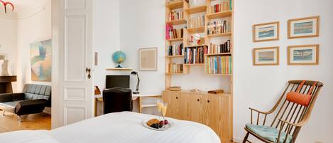 A hotel room with a large bed, a wooden bookshelf, a rocking chair, a desk with a globe, and framed pictures on the wall.