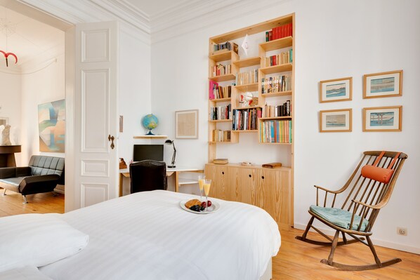 Una habitación de hotel con una cama grande, una estantería de madera, un sillón mecedora, un escritorio con un globo terráqueo y cuadros en la pared.