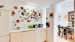 Une cuisine moderne avec des meubles blancs, un four à micro-ondes et une machine à café. Diverses photos et notes sont accrochées au mur, et un coin repas est aménagé avec des chaises et une table.
