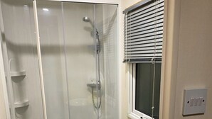 Un baño compacto con ducha de mampara de vidrio, un lavamanos pequeño y una ventana con persianas.