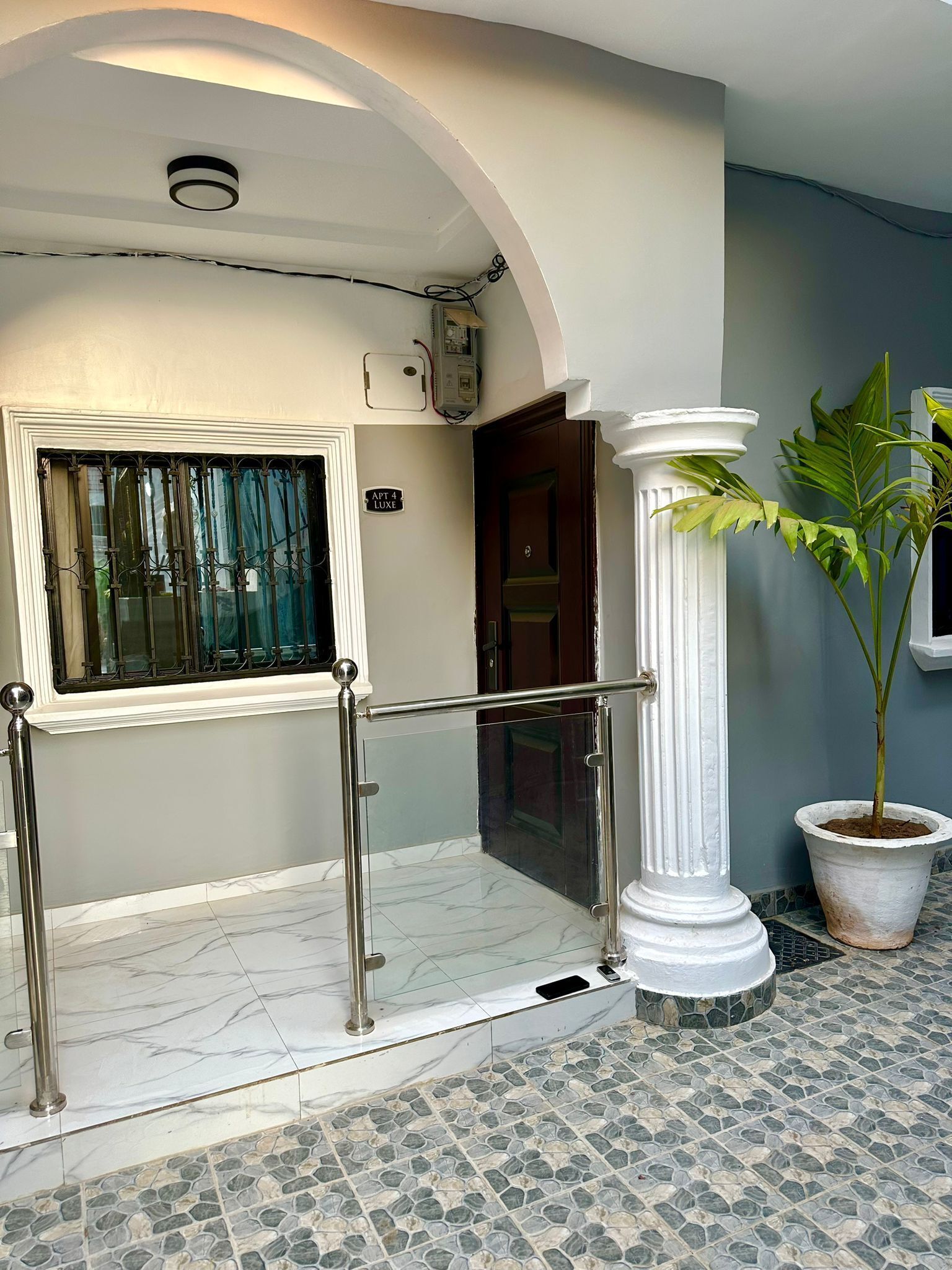 Entrada de hotel com porta de vidro, guarda-corpo de metal, planta em vaso e piso ladrilhado.