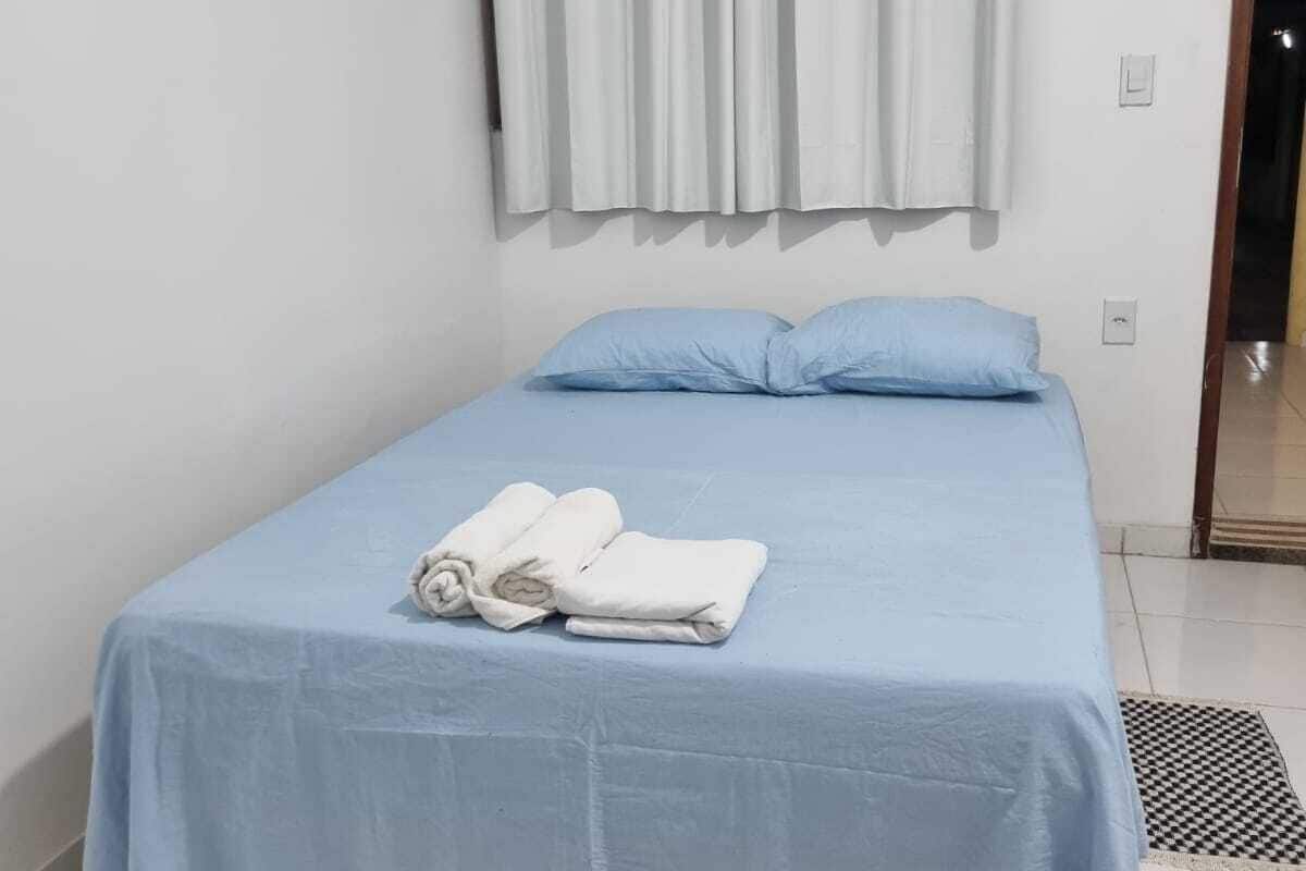 A single bed with a blue bedspread and two pillows, a pair of rolled towels on the bed, and a window with white curtains.