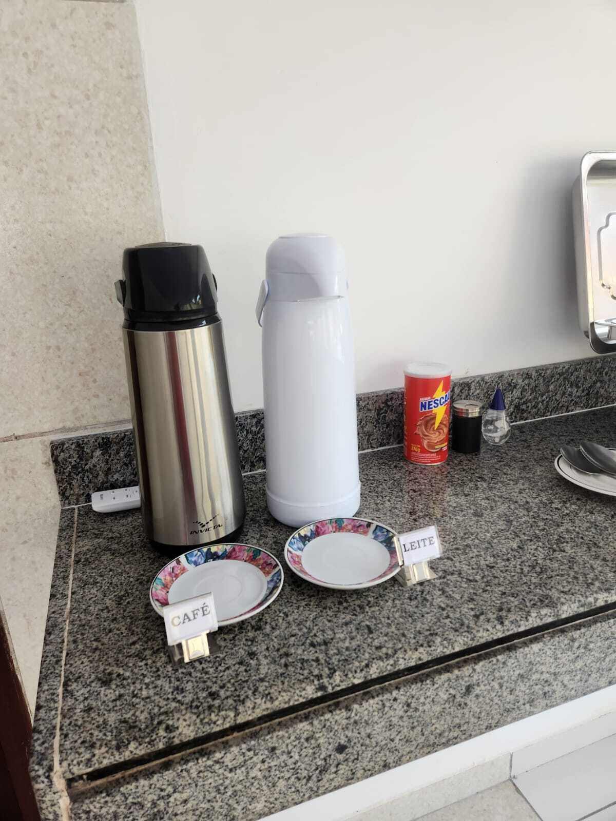Uma bancada de cozinha com uma cafeteira de aço inoxidável, uma chaleira elétrica branca, uma caneca de Nescafé e duas travessas com os dizeres “CAFÉ” e “LEITE”.