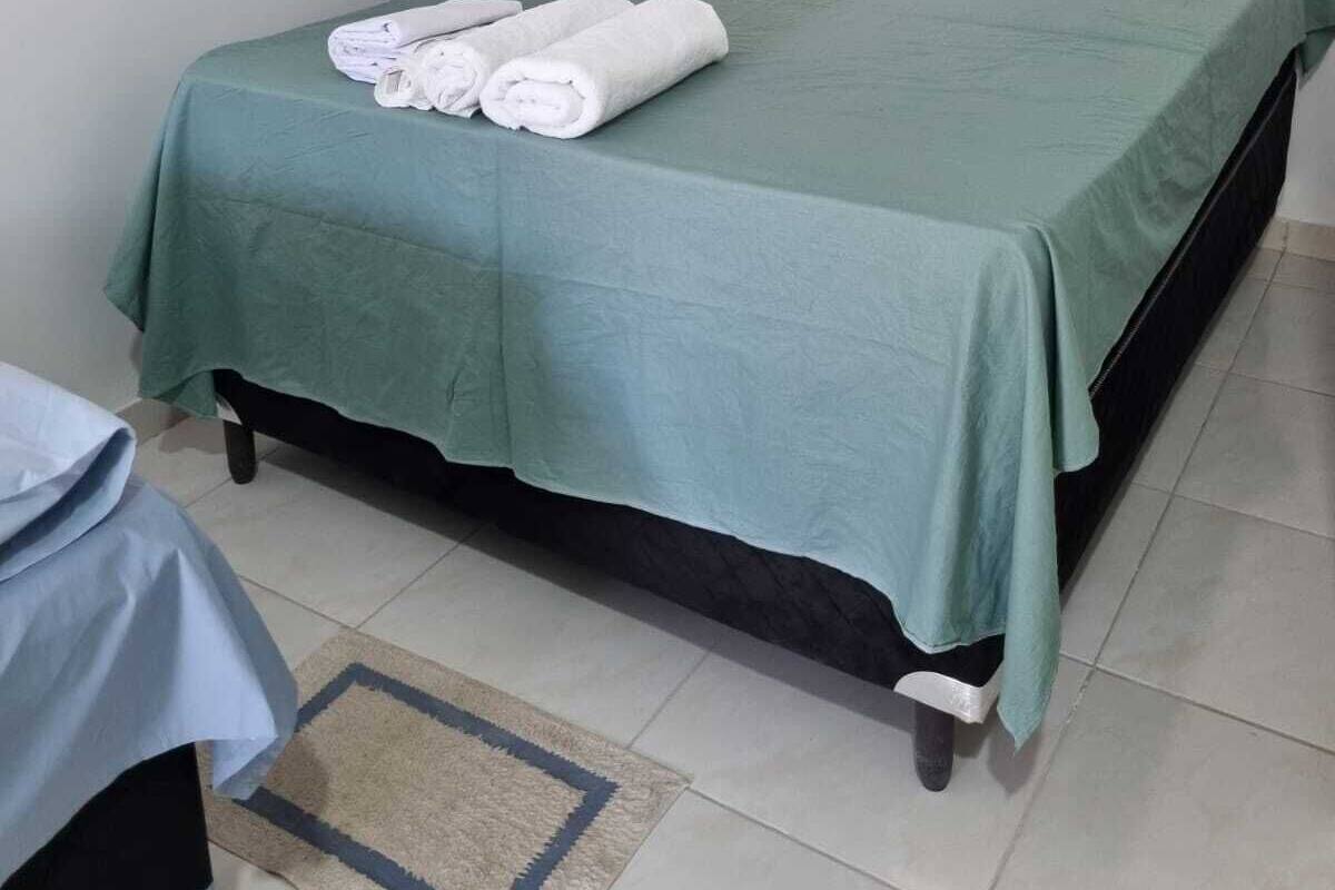 A single bed with a green bedspread and pillows, a rolled white towel on the bed, and a small rug on the floor.