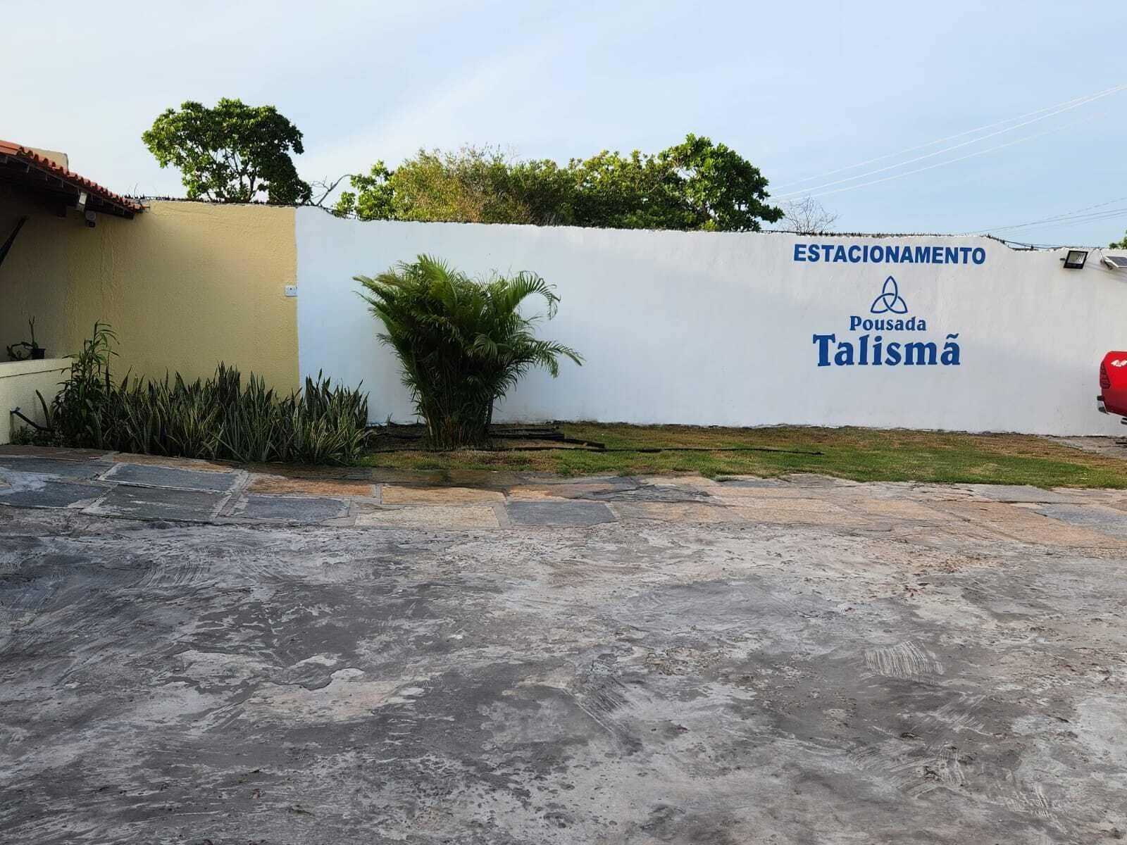 Un área de estacionamiento con un muro blanco que muestra el texto "ESTACIONAMENTO Pousada Talismã".