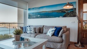 Un salon moderne doté d’une grande fenêtre donnant sur un paysage côtier magnifique, d’un canapé beige agrémenté de coussins à motifs, d’une table basse en verre et d’un lampadaire.