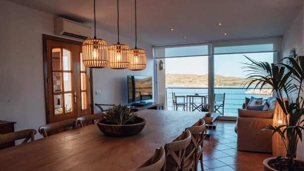 Un vaste espace de salle à manger avec une table et des chaises en bois, une grande fenêtre donnant sur la mer et un coin salon confortable avec un canapé et une plante en pot.