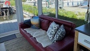 Een rode leren bank met een plaid en kussens, een televisie op een meubelstuk, en uitzicht op boten en een jachthaven door grote ramen.