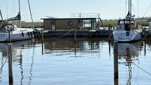 Una marina amb vaixells amarrats, un edifici petit amb una finestra circular i aigua tranquil·la que reflecteix els vaixells.
