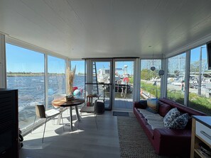 Ein geräumiger Raum mit einem großen Glasfenster zu einem Gewässer, einem roten Sofa, einem kleinen Tisch mit Stühlen und einem Fernseher.