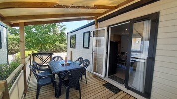 Un porche techado con una mesa y sillas, una puerta corrediza de vidrio que da a un espacio interior, y una terraza de madera.