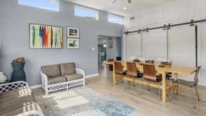 A modern living room with a dining area, featuring a ceiling fan, artwork on the wall, and a view through an open door.