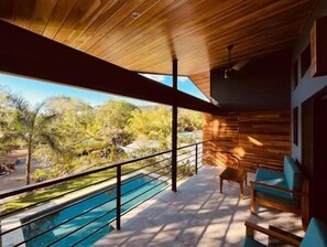 A modern outdoor pool area with a wooden ceiling, a view of a tropical landscape, and a seating area with blue cushions.