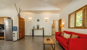 Un salon moderne avec un canapé rouge, un coin cuisine équipé d’un réfrigérateur, d’un distributeur d’eau et d’un meuble en bois. On y trouve également une petite table agrémentée d’objets décoratifs, un luminaire fixé au mur et un escalier menant à un autre niveau.