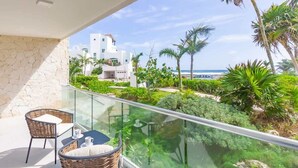 A balcony with wicker furniture, a glass railing, and a view of a white building and palm trees. - Ground-Floor Condo, Beach Club & 5 Rooftop Pools (Akumal)
